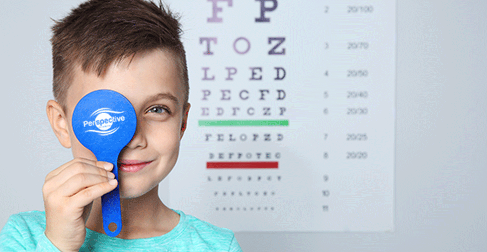 Children's eye exam with a boy holding a Perspective Optometry branded eye occular occluder up to his eye with a snellen visual accuity eye chart behind him.